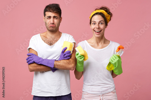Portrait of cheerful attractive young female having excited look, smiling broadly, ready for housework, standing next to her sad unhappy husband who is forced to share household chores with her