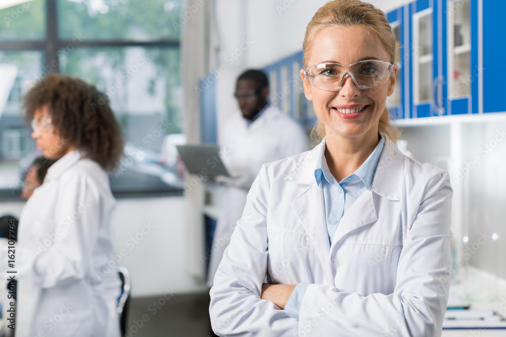 Fototapeta premium Portrait Of Smiling Woman In White Coat And Protective Eyeglasses In Modern Laboratory, Female Scientist Over Busy Researchers Team In Lab