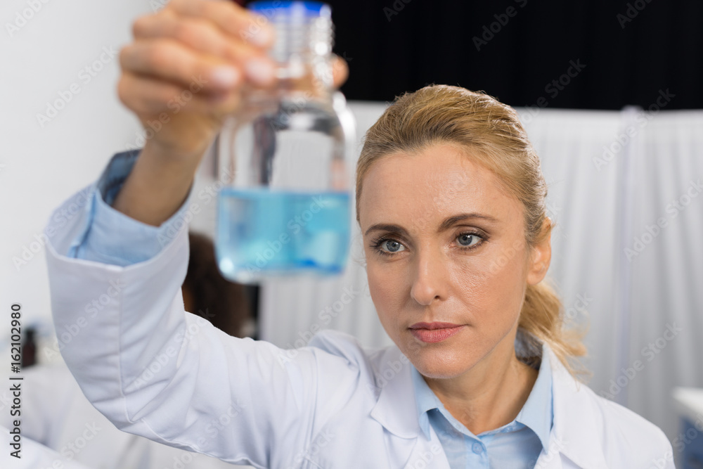 Fototapeta premium Female Scientist Examine Flask With Blue Luquid Working In Modern Laboratory, Attractive Woman Researcher Making Experiment In Lab