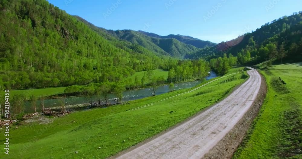 UHD 4K aerial view. Low flight over fresh cold mountain road, river and meadow at sunny summer morning. Green trees and sun rays on horisont. UHD 4K aerial view. Low flight over fresh cold mountain road, river and meadow at sunny summer morning. Green trees and sun rays on horisont.