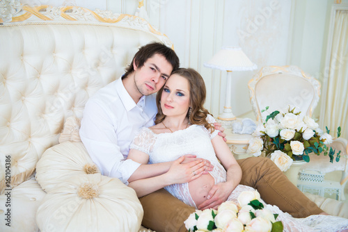 Beautiful couple, pregnant young woman and man, hugging lovingly sitting on the bed, in a home interior.