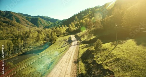 Aerial UHD 4K view. Low air flight over mountain rural dirt road and meadow at sunny summer morning. Near green trees, sun rays and mountain river