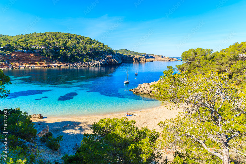 Couple of unidentifed people sunbathing on beautiful beach in Cala Salada famous for its azure crystal clear sea water, Ibiza island, Spain