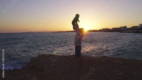 Father holds his baby boy over the head. Silhouettes against sea sunset. Slow motion shot