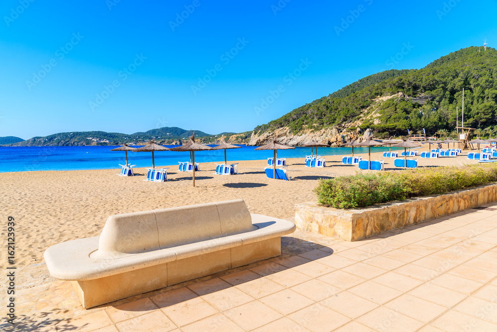Sandy beach with umbrellas and sunbeds in Cala San Vicente bay on sunny summer day, Ibiza island, Spain