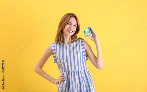 Happy young woman holding piggy bank on color background