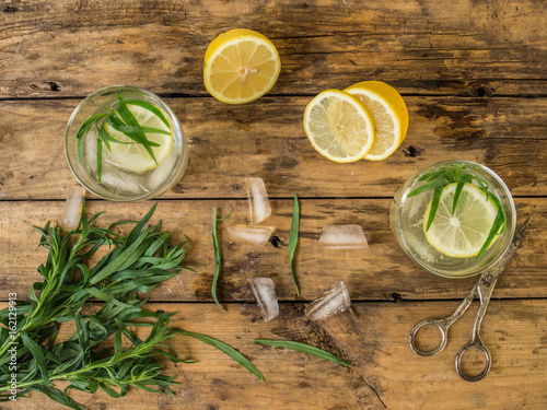 Homemade lemonade tarragon in a glass jug and in two glasses served with lemon wedges and tarragon sprigs on an old wooden background. Top view