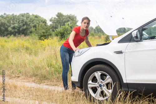 Young woman looking under the hood of overheated car