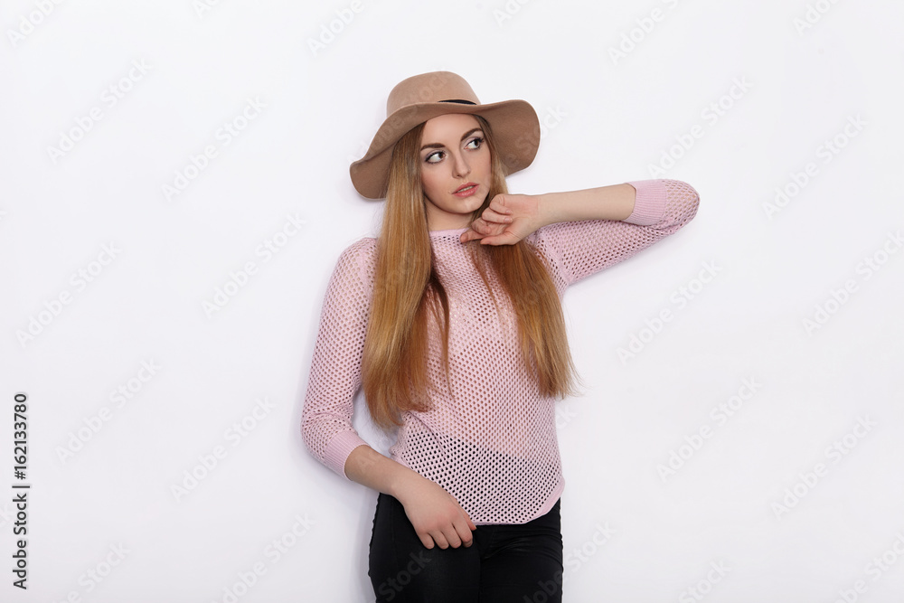 Cute and stylish beautiful young blonde woman in trendy clothing practicing model poses and looking away with smile while standing against white wall background Cute and stylish beautiful young blonde woman in trendy clothing practicing model poses and looking away with smile while standing against white wall background