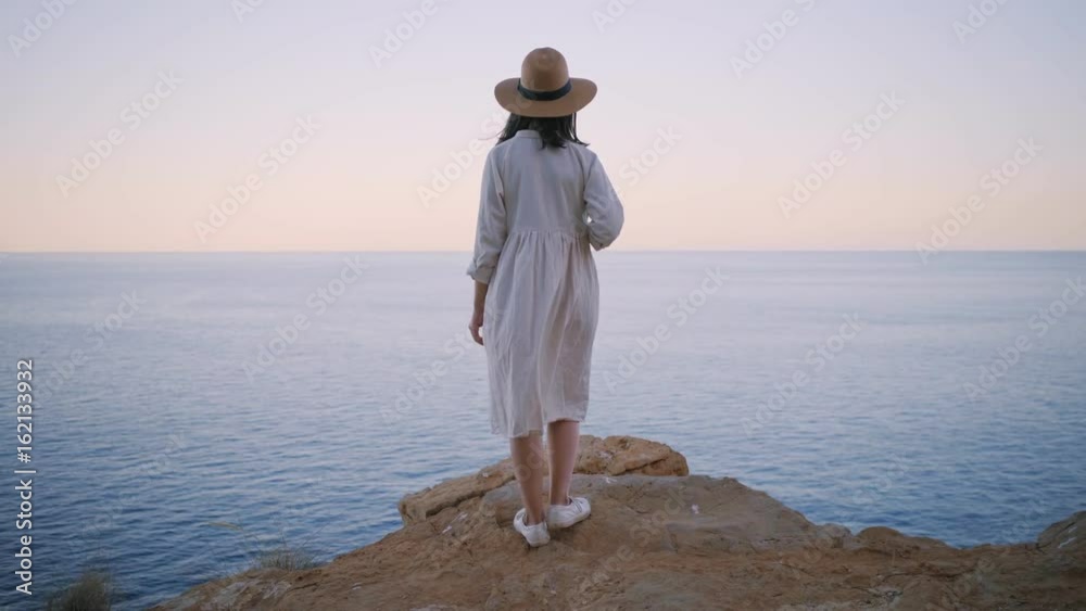Rear view on shy and dreamy amish girl in simple white cotton dress and straw hat, standing on the edge of rock cliff and looking far away in ocean sea, waiting someone. Wind slightly swings her dress