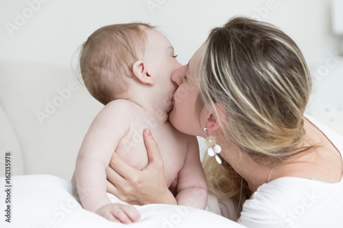 Portrait of happy laughing mother kissing her baby boy on bed