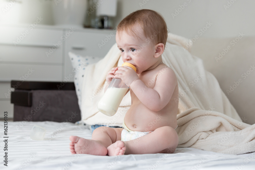 Beautiful 9 months old baby boy sitting on bed and drinking milk Beautiful 9 months old baby boy sitting on bed and drinking milk