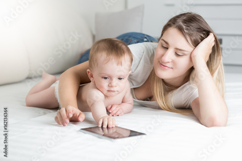 Beautiful smiling mother and 10 months old baby boy lying on bed with tablet PC