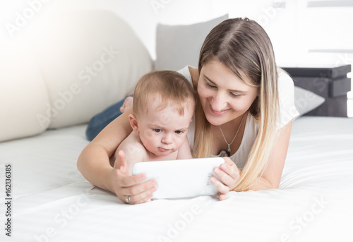 Beautiful young mother lying with her baby on bed and using tablet