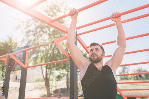 Closeup of strong athlete doing pull-up on horizontal bar.Mans fitness with blue sky in the background and open space around him. Young man with sportive clothes in the city