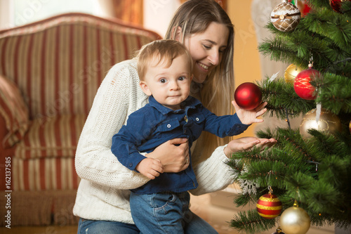 Mother with her 10 months old baby boy decorating Christmas tree at living rom