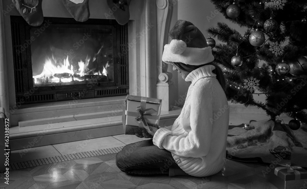 Black and white image of girl holding gift box and sitting next fireplace