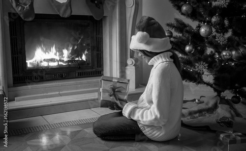 Black and white image of girl holding gift box and sitting next fireplace