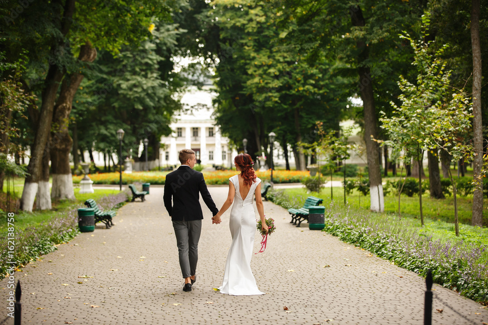 Fototapeta premium Stylish bride and groom holding hands and walking in park near place of wedding ceremony