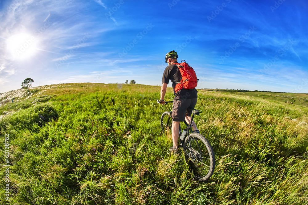 Young cyclist riding mountain bicycle through green meadow against beautiful sky.