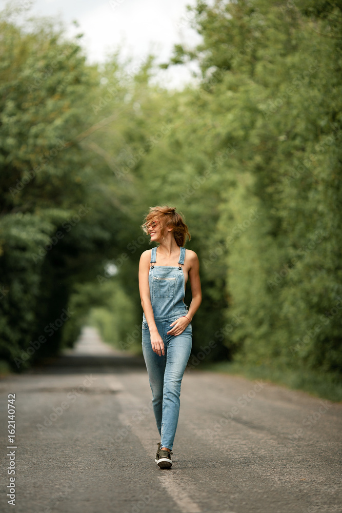 Young woman in denim overalls and sunglasses goes along forest road. Young woman in denim overalls and sunglasses goes along forest road.