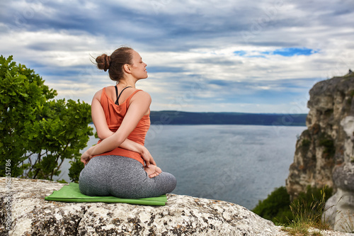 Rear view of young woman in yoga pose sitting on the rock above river.
