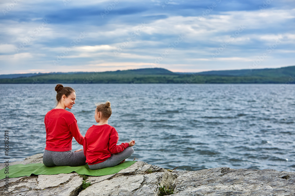 Rear view of the mother and daughter are practicing yoga on the rock near river. Rear view of the mother and daughter are practicing yoga on the rock near river.