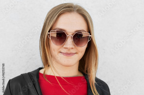 Close up of young female wearing trendy sunglasses, red sweater and black leather coat having pleased expression isolated over white background. Stylish adorable woman posing into camera outside