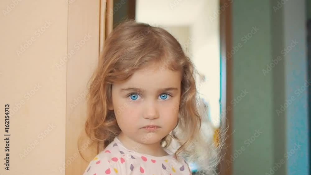 Little girl is blonde with blue eyes stands near the wall in the house