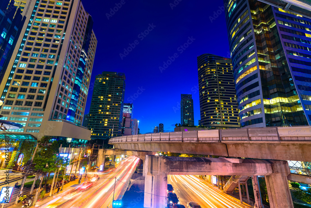 Fototapeta premium Photo of commercial office buildings exterior. Night view at bottom skyscrapers with light of traffic rush, long exposur in Bangkok