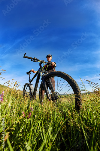 Young cyclist in the helmet in the green meadow on a background with blue sky. © Aleksey