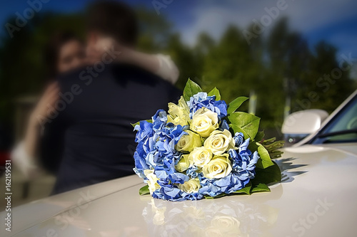 Bridal bouquet on a wedding car. In the background hugging the bride and groom blurry