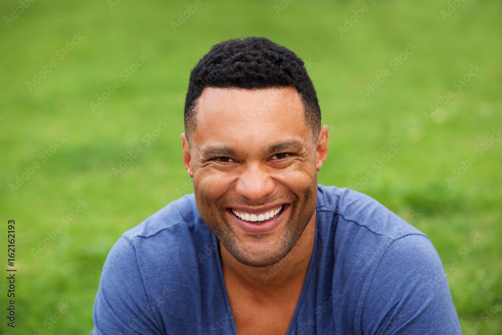 Close up smiling young man sitting outdoors against green grass Close up smiling young man sitting outdoors against green grass