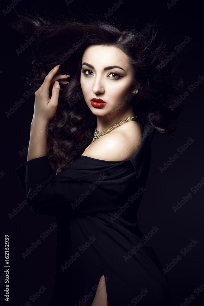 Portrait of glam dark-haired model with perfect make up and flying wavy silky hair wearing golden luxurious necklace and black side split slit top. Isolated on black background. Studio shot