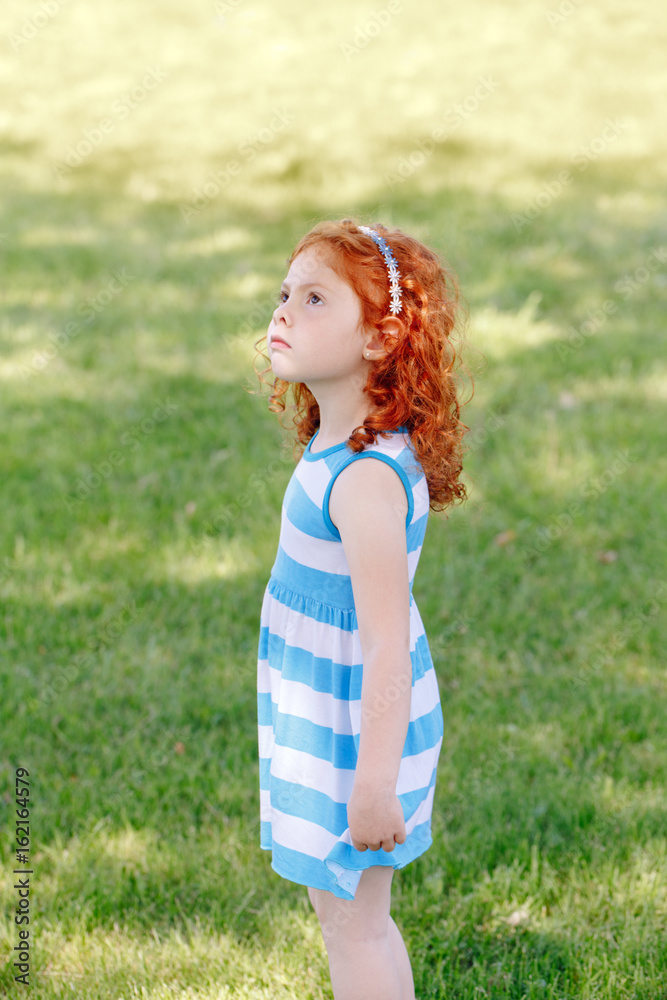 Portrait of cute adorable little red-haired Caucasian girl child in blue dress standing in field meadow park outside, looking up in the air, having fun, happy lifestyle childhood concept Portrait of cute adorable little red-haired Caucasian girl child in blue dress standing in field meadow park outside, looking up in the air, having fun, happy lifestyle childhood concept