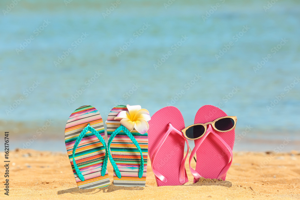 Fototapeta premium Colorful flip-flops, sunglasses and flower on sand at sea shore. Vacation concept