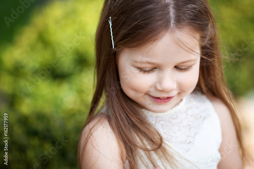 Beautiful smiling little girl in cream dress, against green of summer park