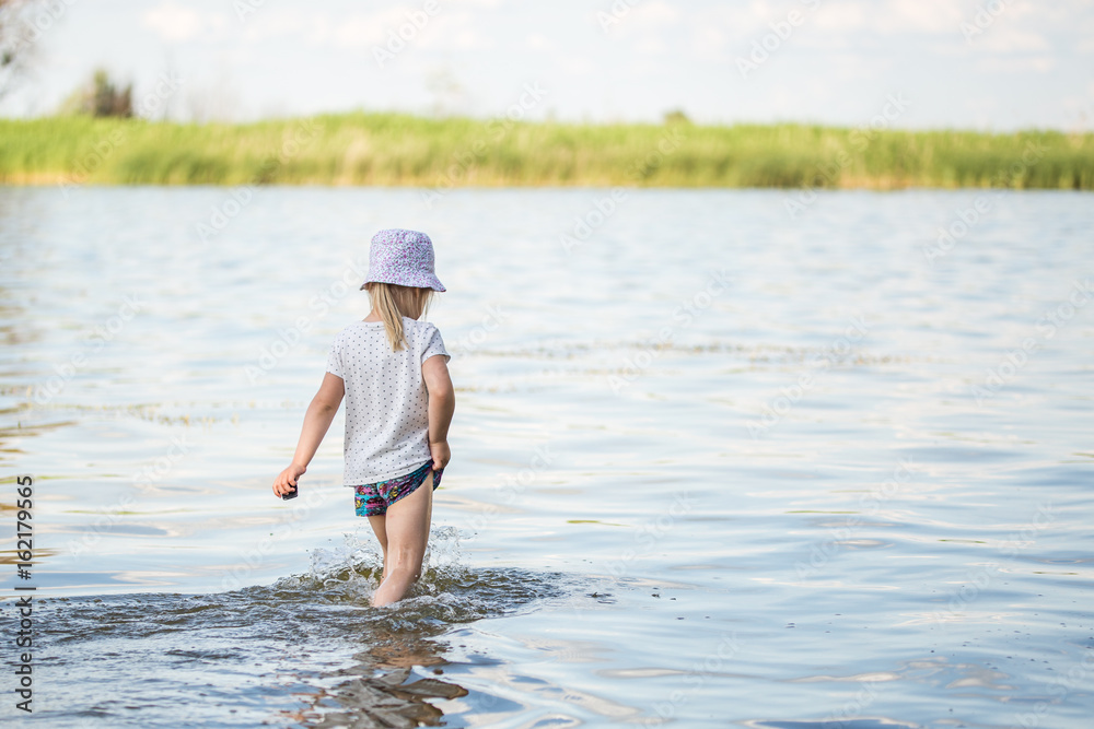 Cute little blond girl walking along the beach of a lake or river. Back view