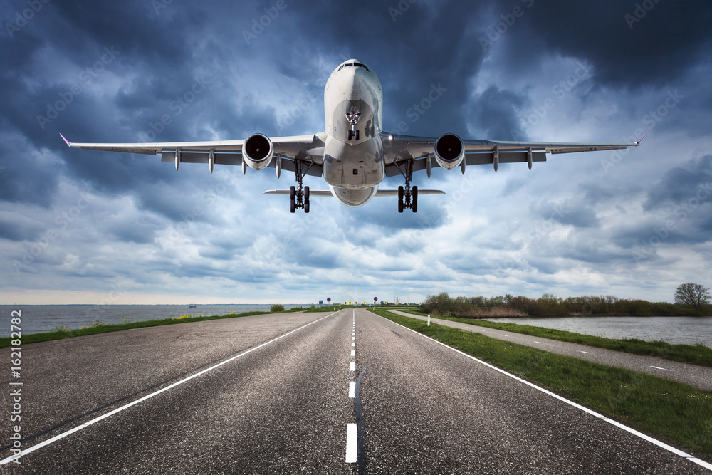 Fototapeta premium Samolot i droga. Krajobraz z dużym białym samolotem pasażerskim leci w pochmurne niebo nad drogą asfaltową. Podróż. Samolot pasażerski ląduje na pasie startowym. Samolot komercyjny i autostrada
