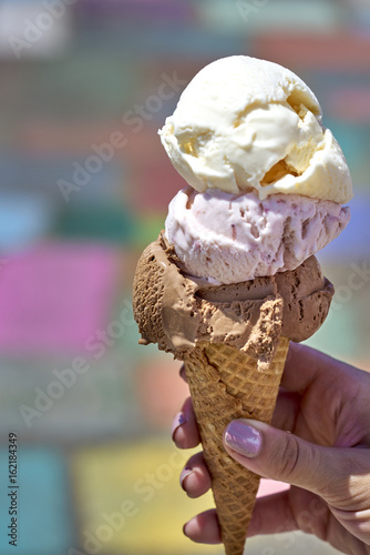 Chocolate, vanilla and berry ice cream scoops in waffle cone held by female hand against colorful background