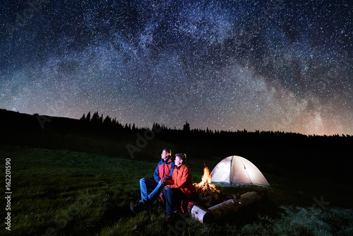 Night camping. Man and woman tourists have a rest at a campfire near illuminated tent under beautiful night sky full of stars and milky way. Low light