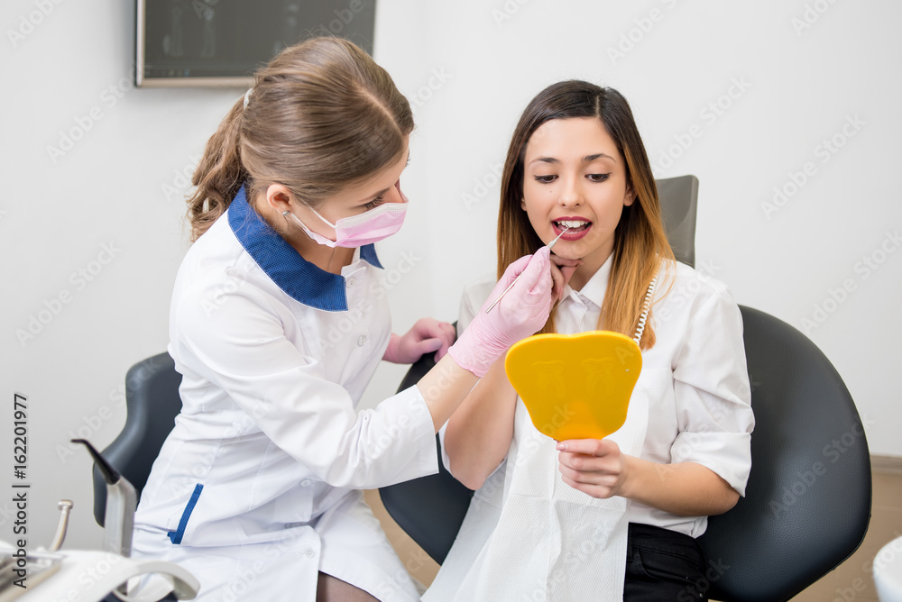 Beautiful woman patient and female dentist with probe are checking teeth at mirror after dental treatment in modern dental clinic. Dentistry Beautiful woman patient and female dentist with probe are checking teeth at mirror after dental treatment in modern dental clinic. Dentistry
