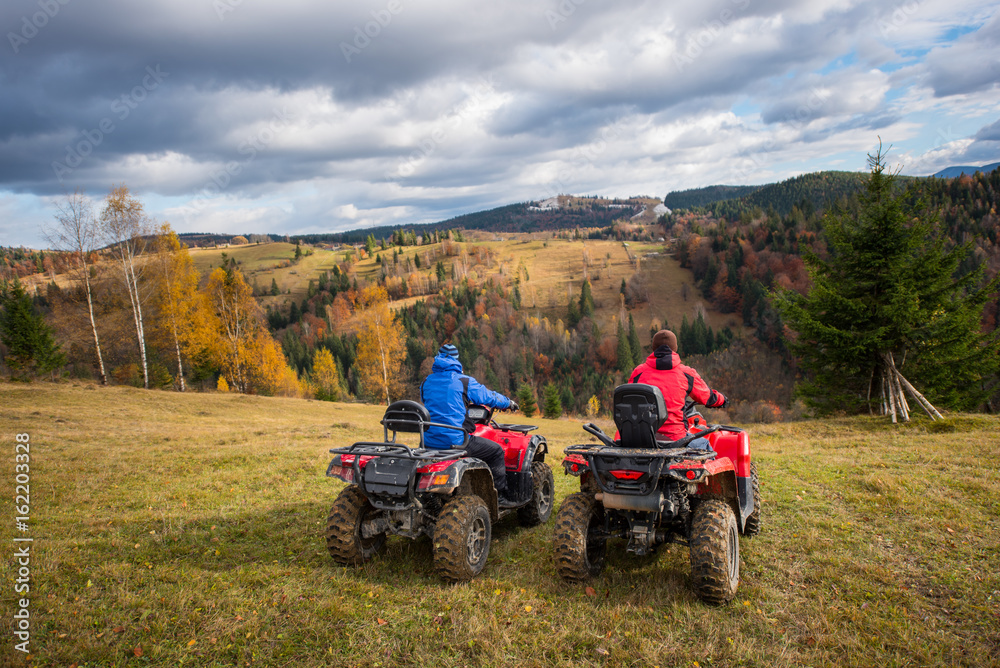 Rear view of two men sitting on quad bikes enjoying beautiful landscape of mountains and colorful forest under the sky with cumulus clouds in autumn Rear view of two men sitting on quad bikes enjoying beautiful landscape of mountains and colorful forest under the sky with cumulus clouds in autumn