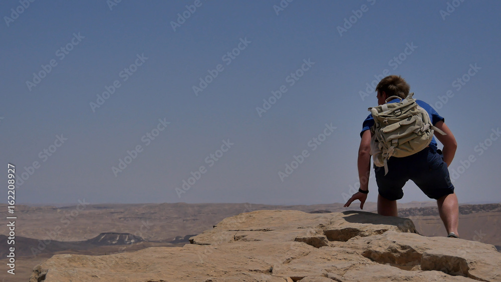 Young man with backpack sitting on cliff's edge and looking at the desert