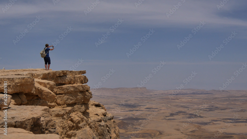 Young man standing on cliff edge and taking panoramic photo of the desert on his phone
