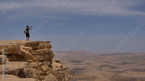 Young man standing on cliff edge and taking panoramic photo of the desert on his phone