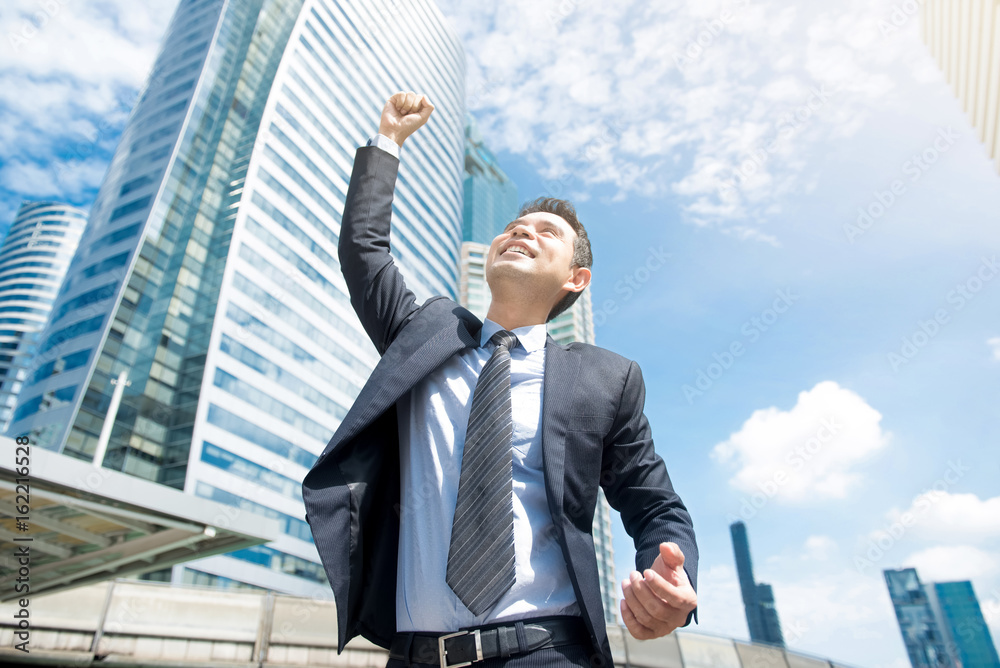 Businessman smiling and raising his fist in the air, with office building background Businessman smiling and raising his fist in the air, with office building background