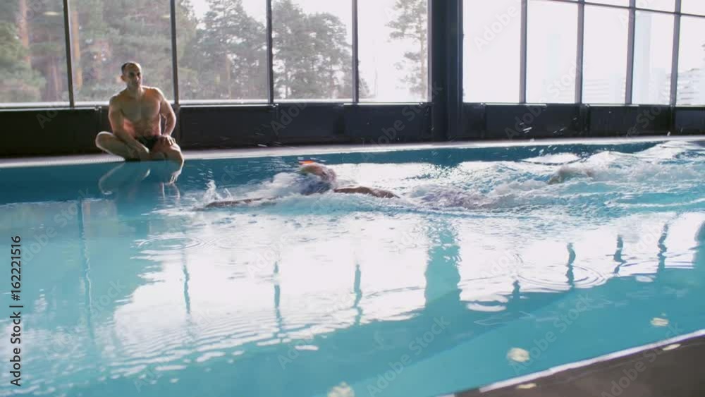 PAN with slow motion of female athlete in goggles and cap swimming with front crawl in indoor pool with blue water while muscular male coach sitting on edge and watching her technique PAN with slow motion of female athlete in goggles and cap swimming with front crawl in indoor pool with blue water while muscular male coach sitting on edge and watching her technique