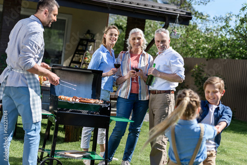smiling man cooking food on barbecue with family enjoying weekend near by