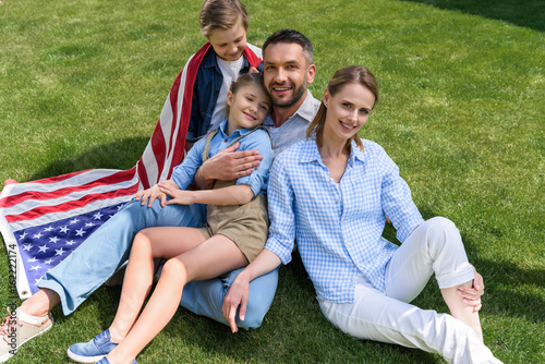 Happy parents and children relaxing on grass with american flag, celebrating 4th july - Independence Day